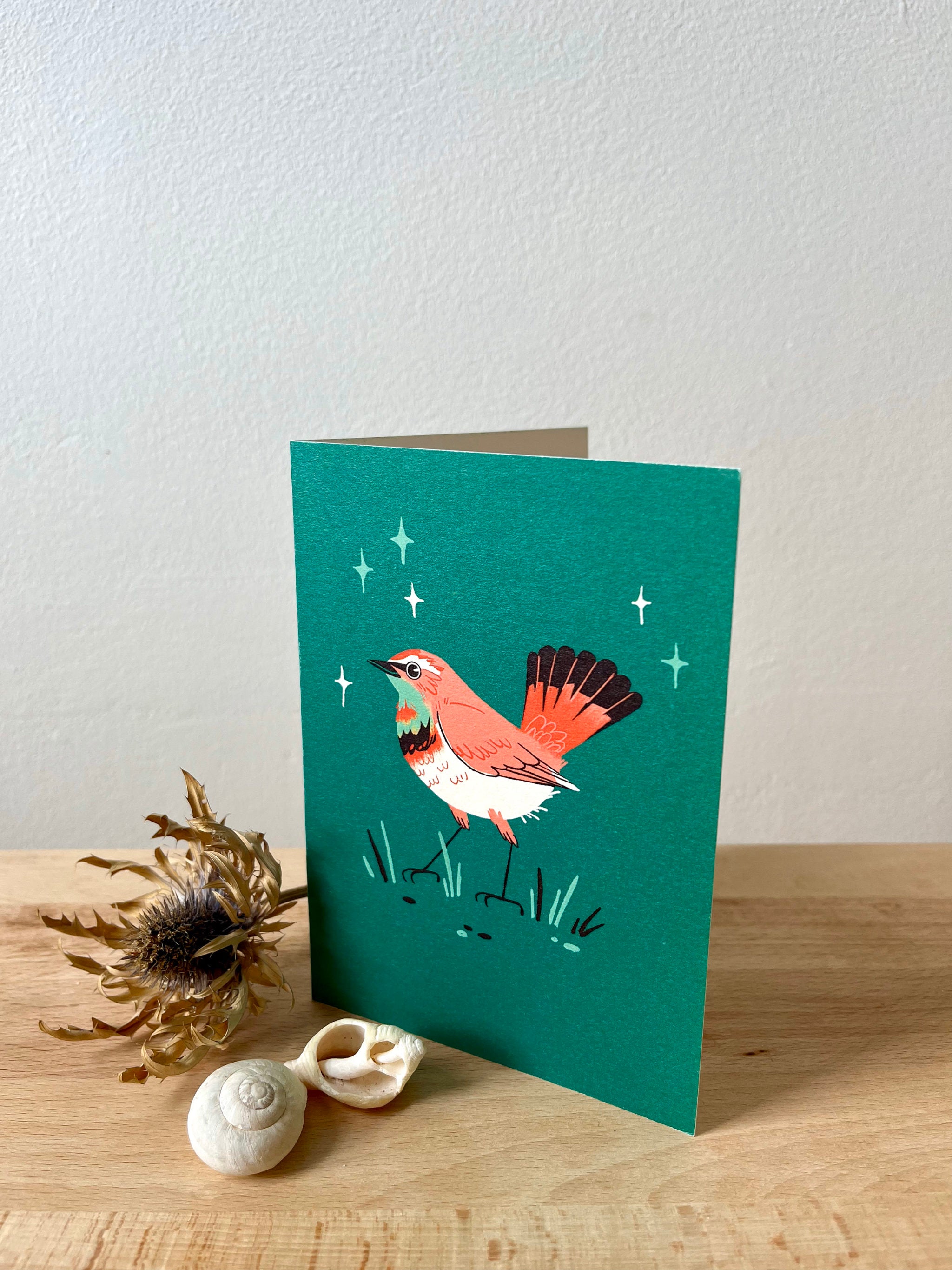 Bluethroat greetings card -- greeting card with a bird illustration on a teal background, placed on a wooden surface with decorative elements.