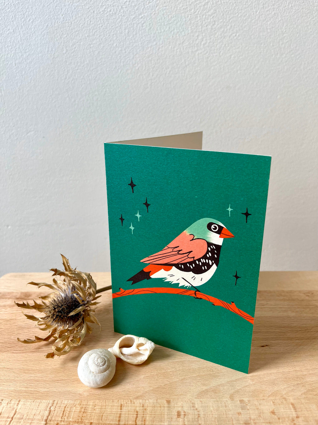 Diamond Firetail greetings card -- greeting card with a bird illustration on a teal background, placed on a wooden surface with decorative elements.