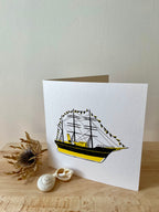 Discovery greetings card -- greeting card with a ship design on a wooden surface with shells and dried plant.