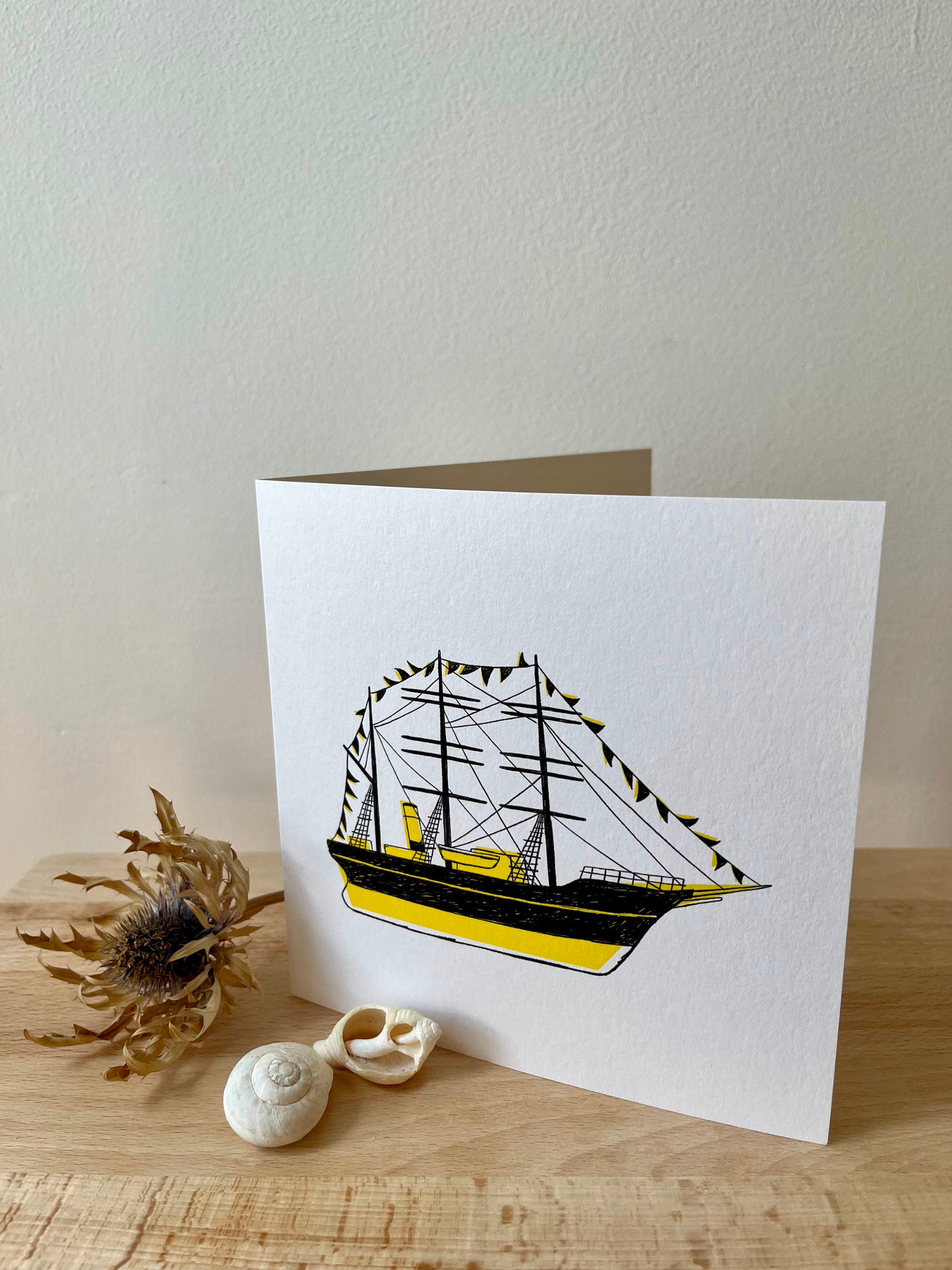 Discovery greetings card -- greeting card with a ship design on a wooden surface with shells and dried plant.