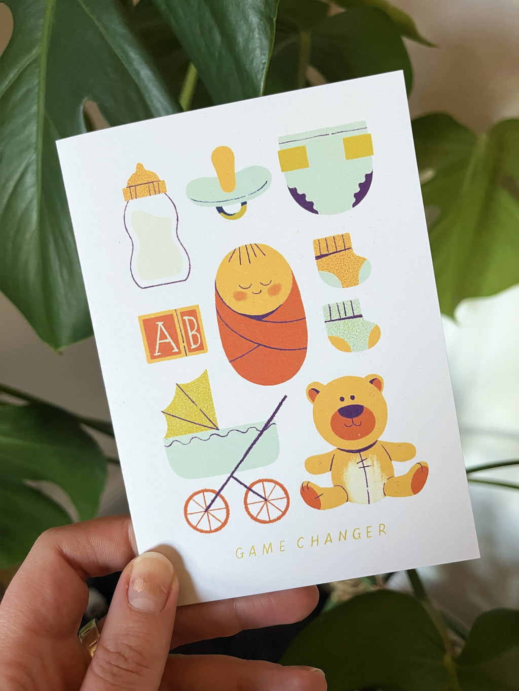 Game Changer greetings card -- card with baby-themed illustrations held by a hand against a leafy background.