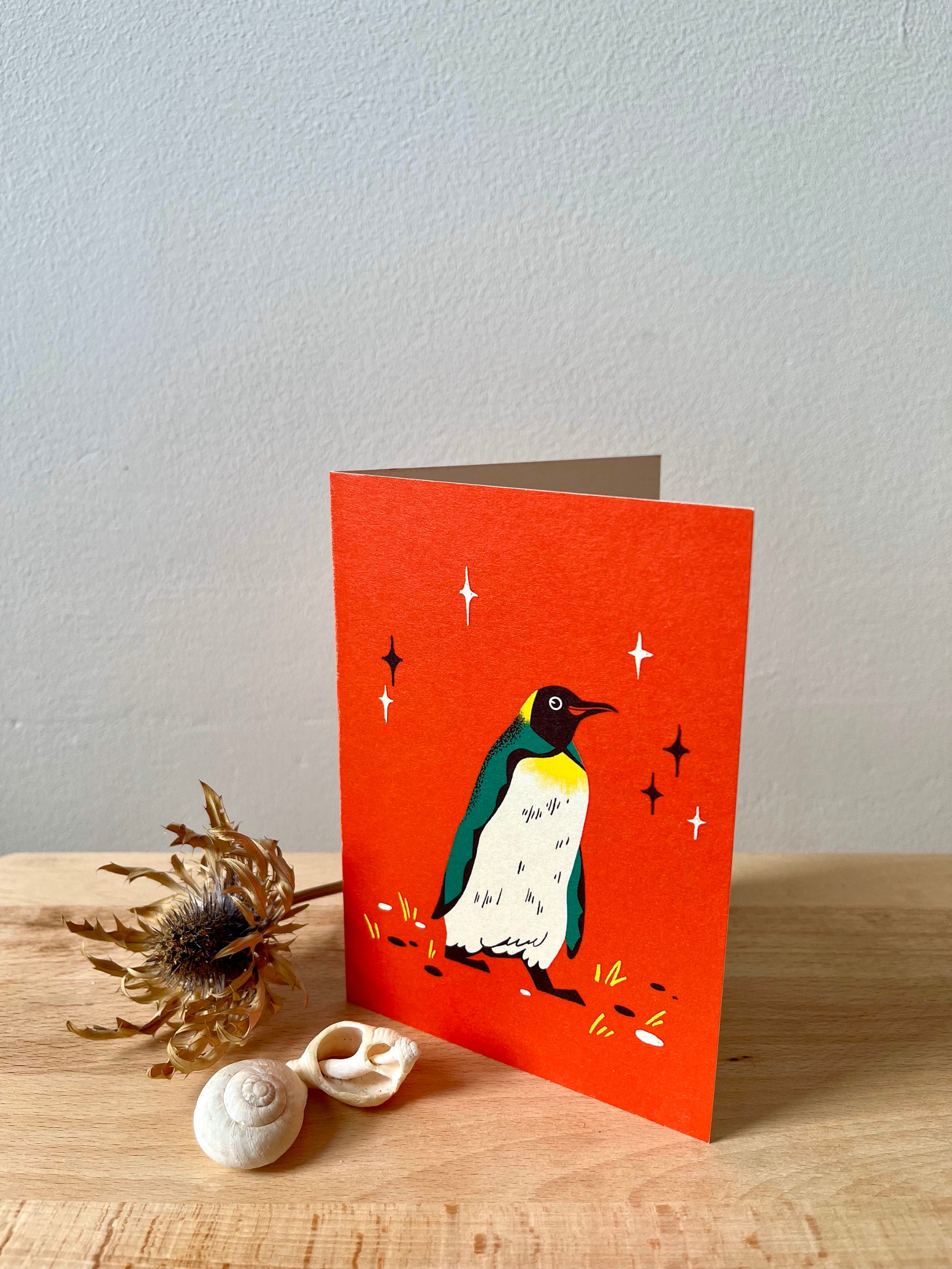 King Penguin greetings card -- greeting card with a penguin illustration on a red background, placed on a wooden surface with decorative elements.