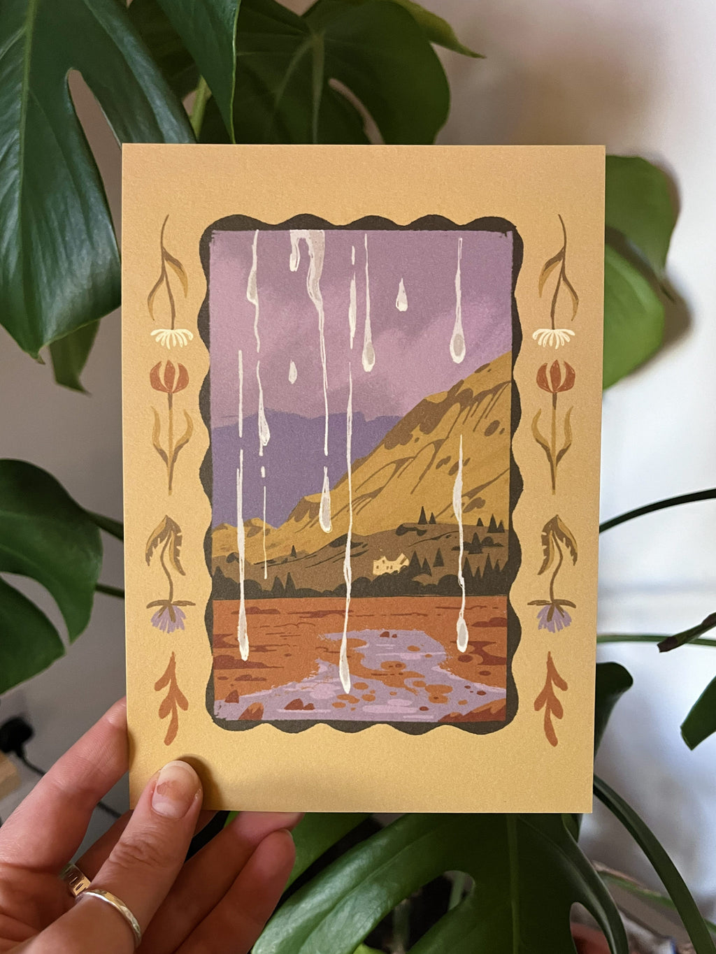 Rainy Bus Journey print -- hand holding a card with a scenic illustration of mountains and water, framed by decorative elements.