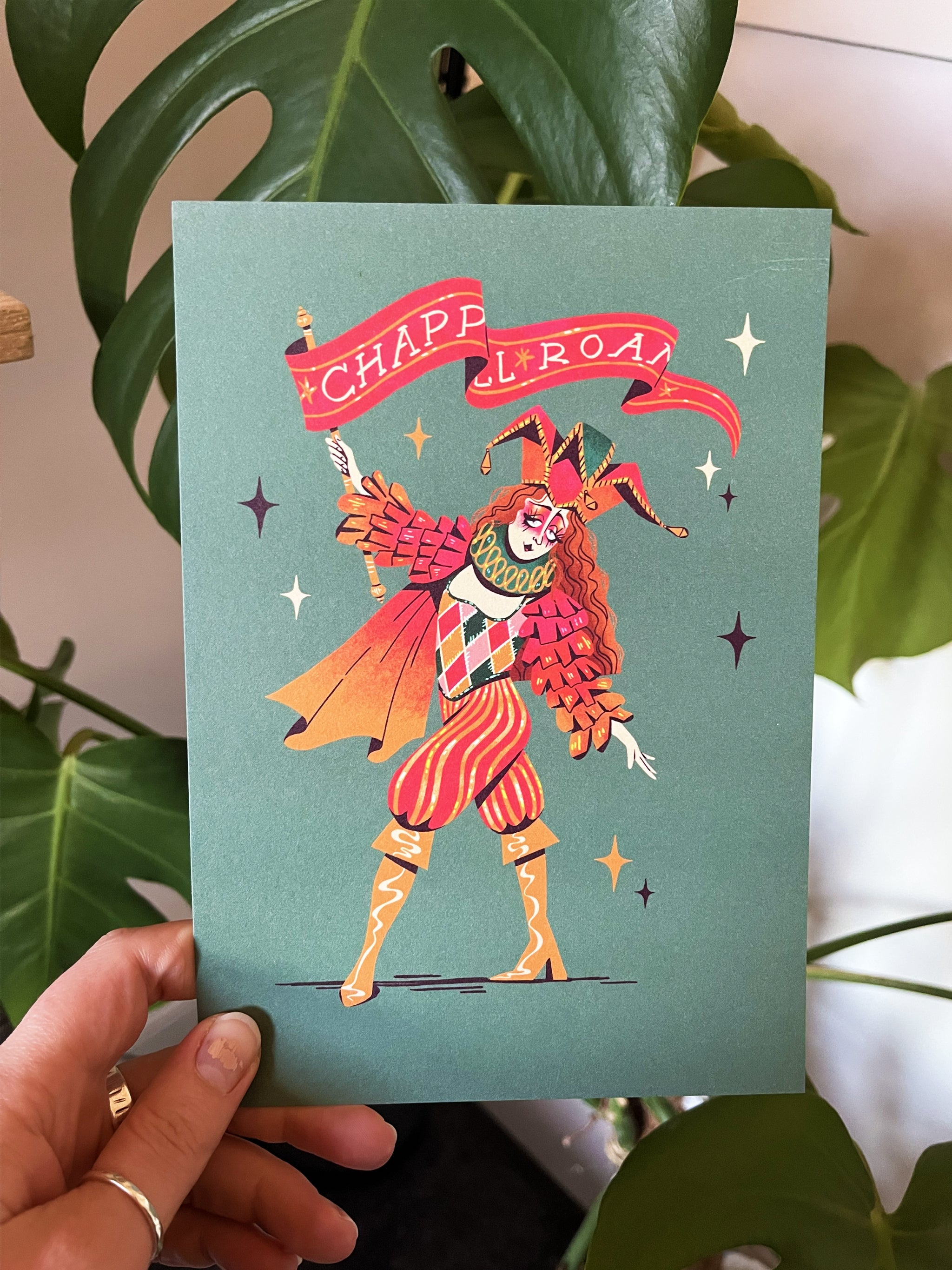 Chappell Roan print -- greeting card with a jester design held by a hand in front of green leaves.