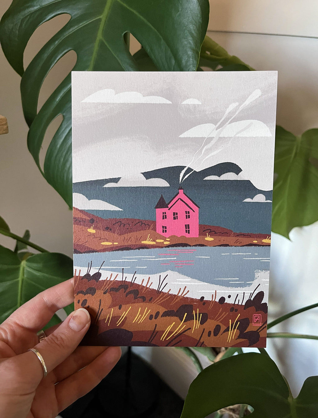 Pink House greetings card -- illustrated card with a pink house on a hill, held by a hand against a plant background.