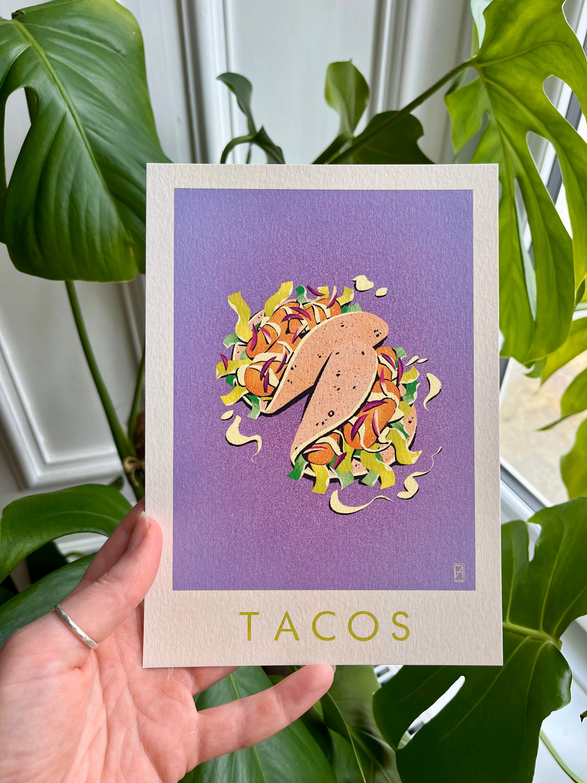 Taco print -- hand-held A5 card with a taco illustration on a purple background held in front of green plants.