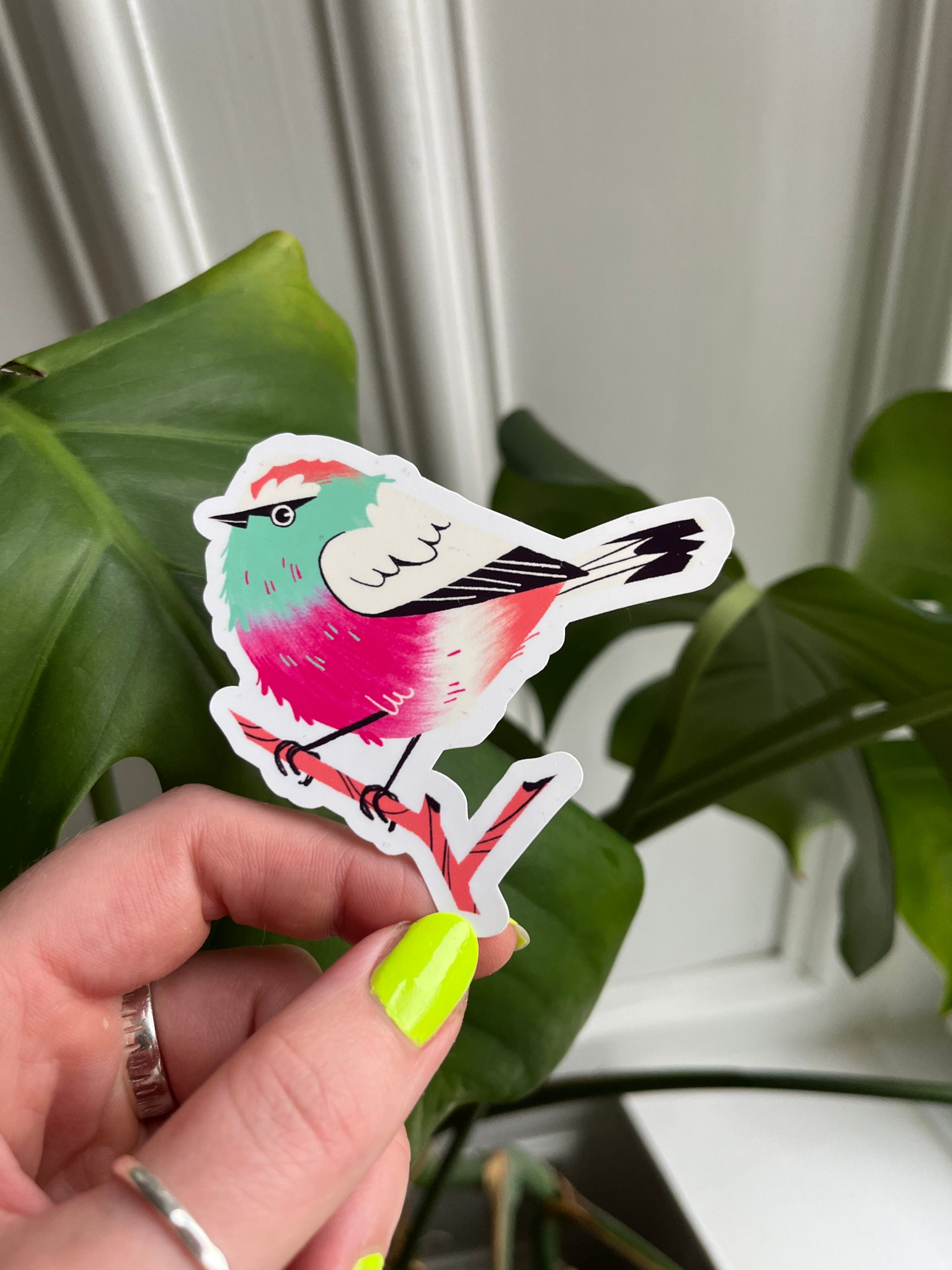 White-browed Tit Warbler -- colorful bird-shaped sticker held by a hand with green nail polish against a leafy background.